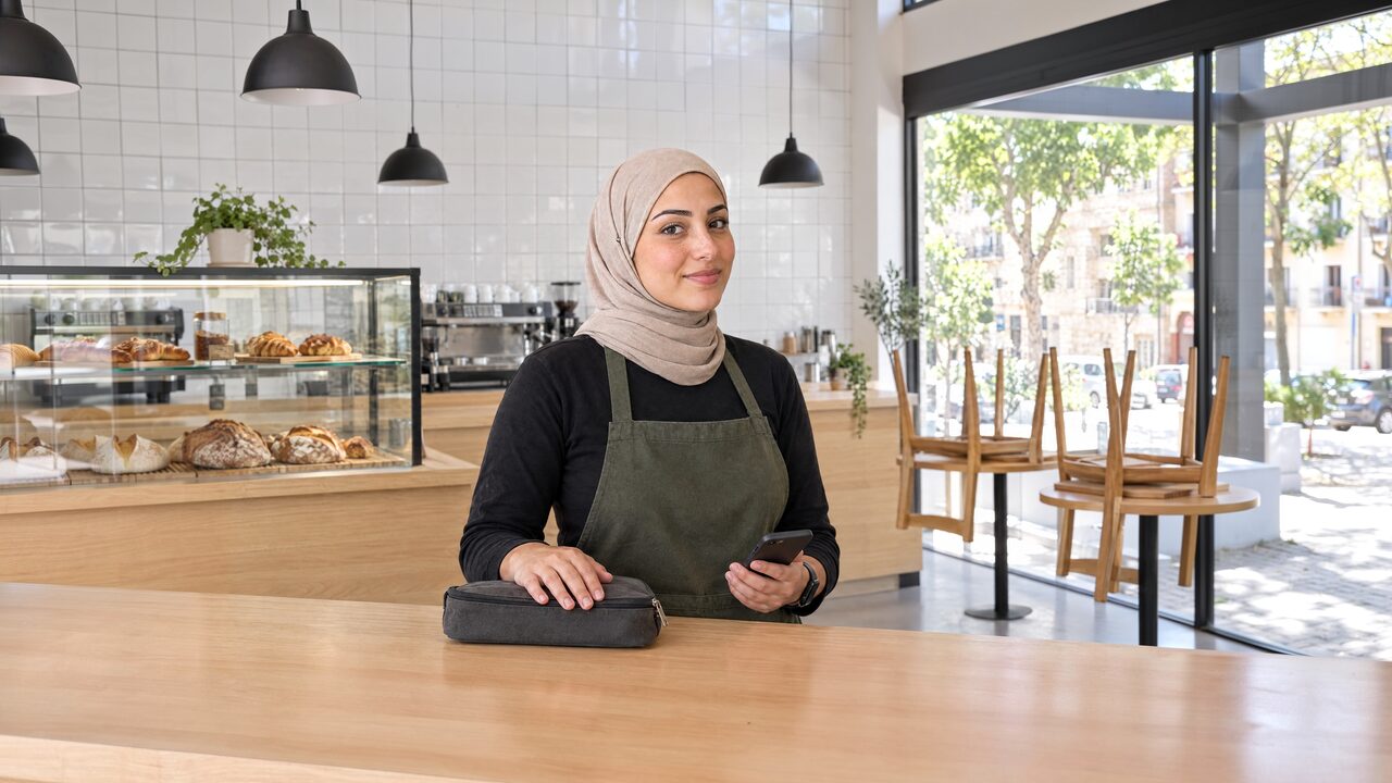 אחראית משמרת בחיג'אב בז' וסינר ירוק זית מאחורי דלפק עץ בבית קפה מואר, כף ידה על נרתיק מזומן סגור